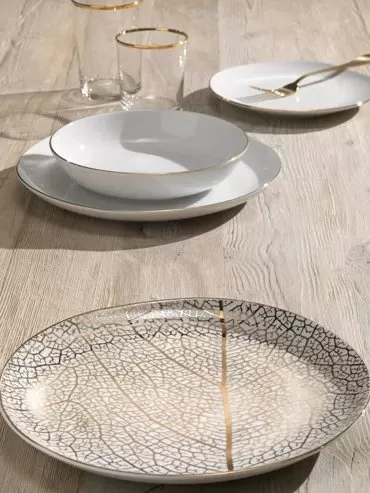 Piatti bianchi con il bordo oro e un piatto da portata decorato a foglia, con bicchieri su un tavolo in legno
